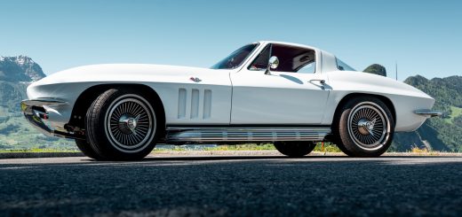 Low side view of a white classic sports coupe on asphalt, with mountains and clear blue sky behind it.